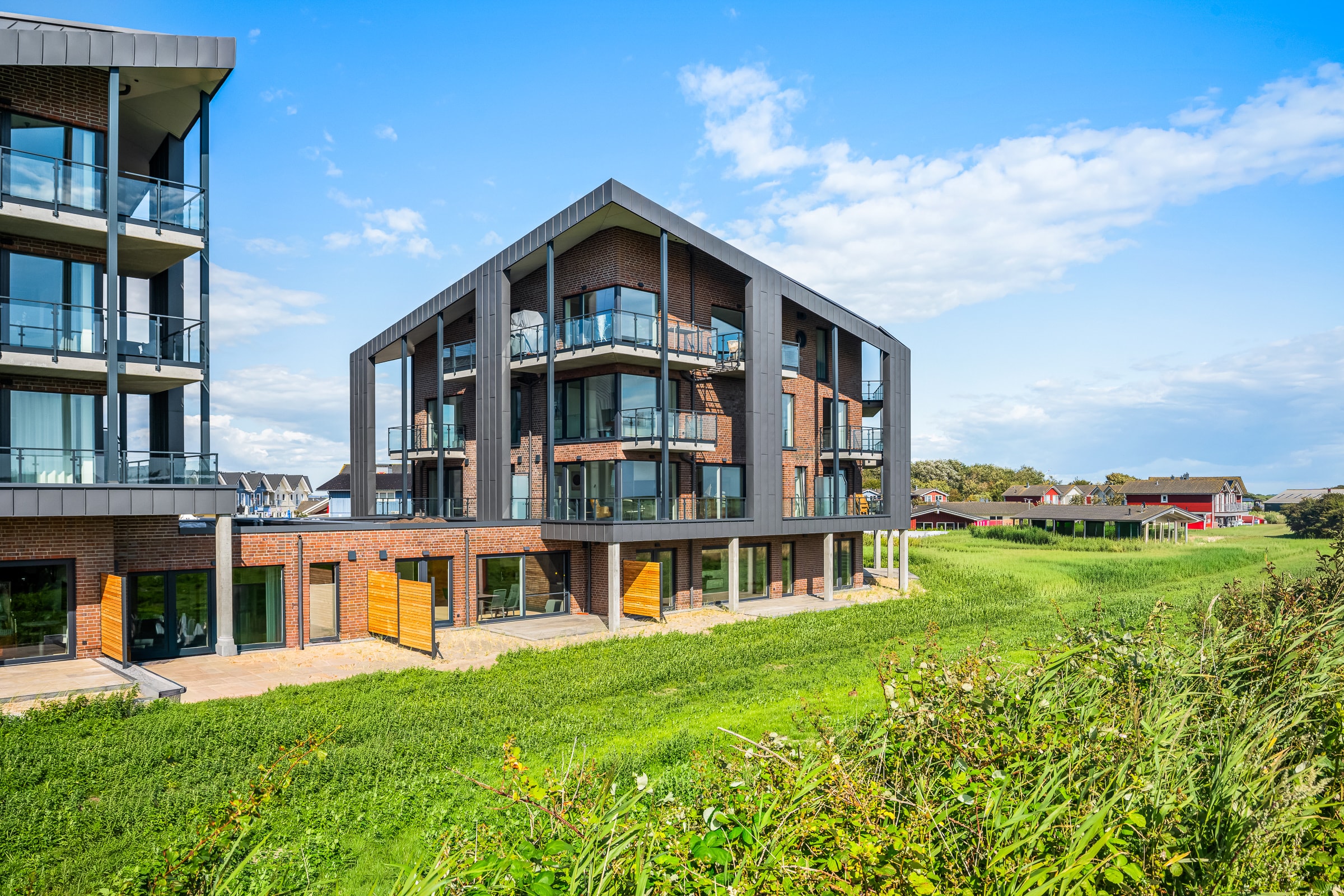 Nowy apartamentowiec The View w Dagebüll: elewacja klinkierowa z blachą na rąbek stojący, balkony ze szklanymi balustradami, z przodu zielone nadmorskie łąki