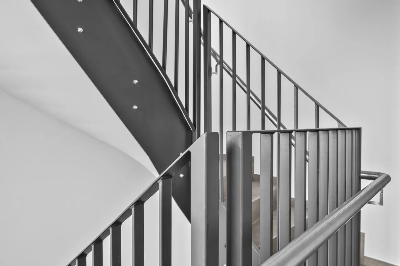 Nawrót balustrady przy spoczniku: słupki, pionowe pręty i gięta poręcz nierdzewna przed białą ścianą